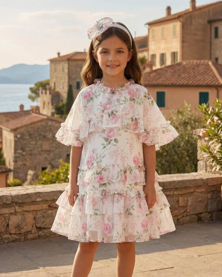 Young girl in a floral dress standing in an outdoor setting with buildings and water in the background.