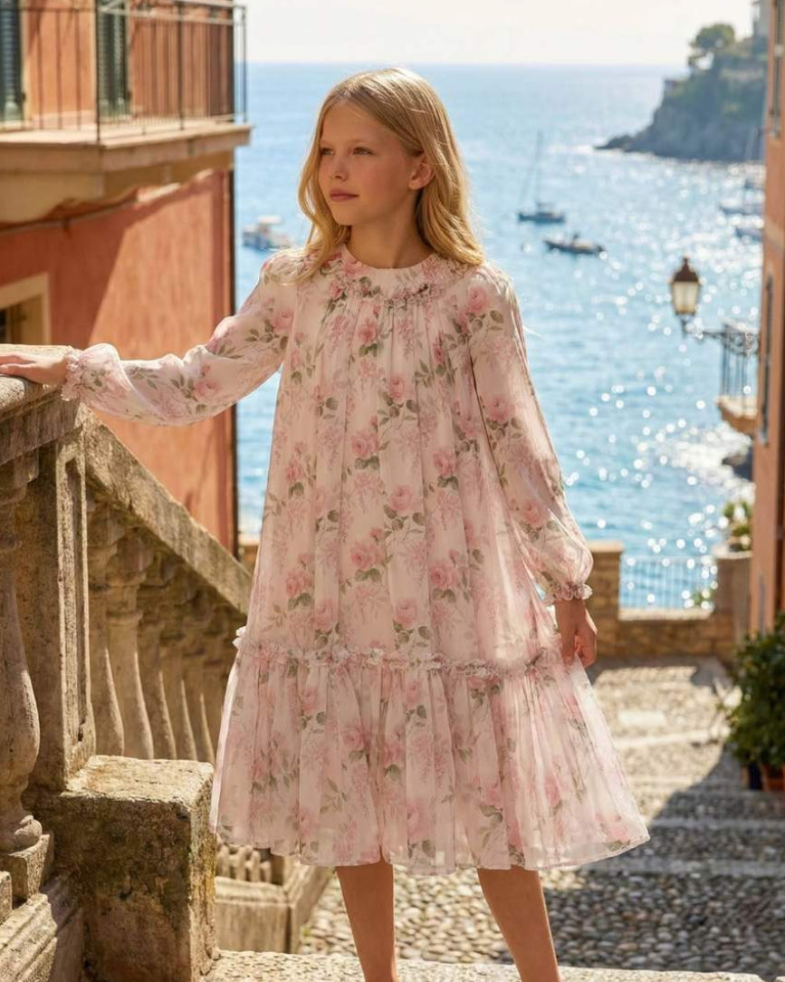 Woman in a floral dress standing on a balcony overlooking the ocean.